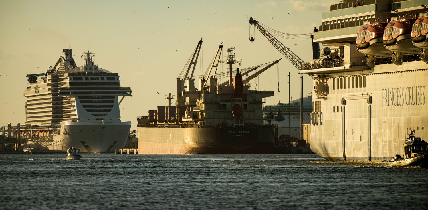 A panoramic view of industrial port facilities and cargo ships along a waterfront during sunset.