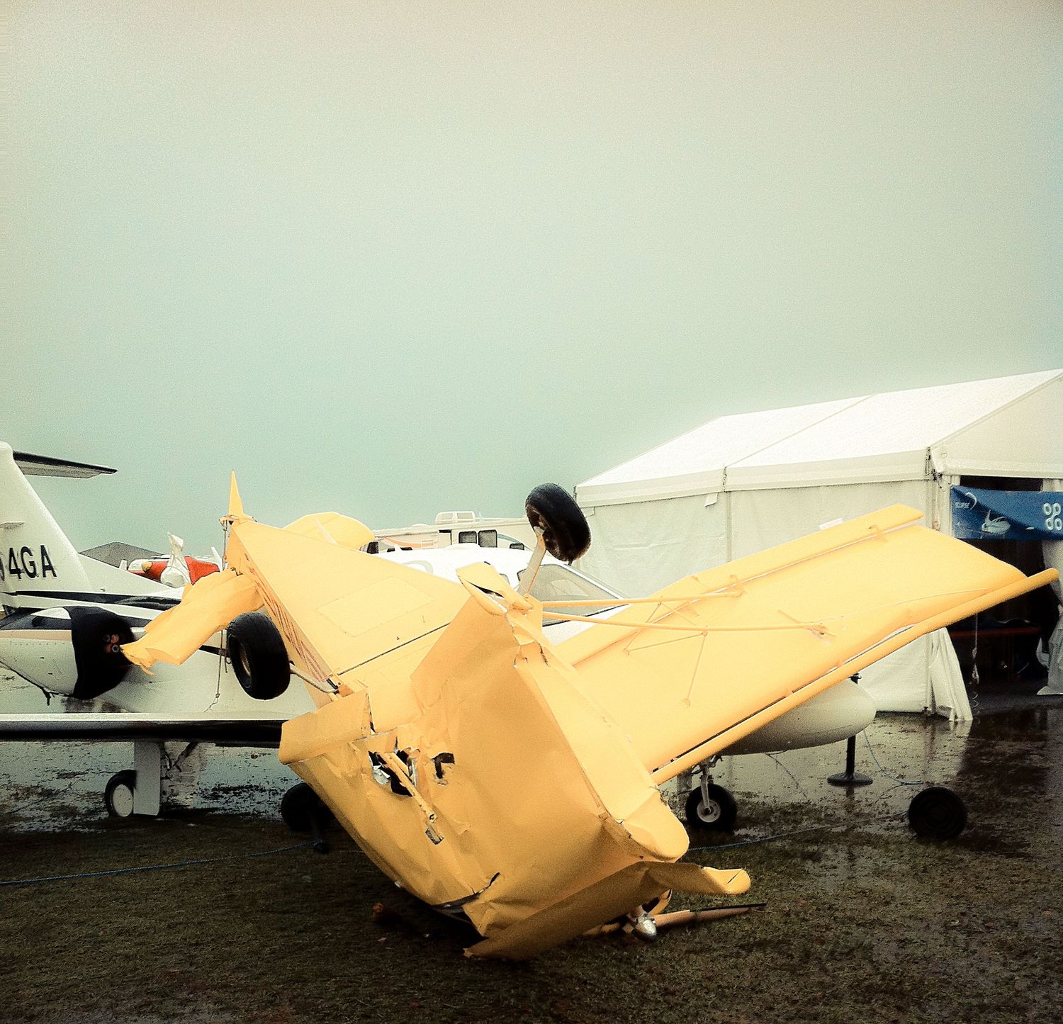 A small yellow aircraft lies overturned on its back in muddy terrain with other planes visible in the background.