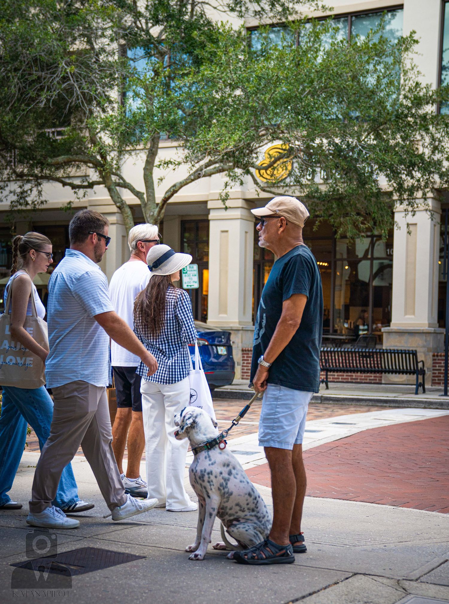People gather on a brick sidewalk outside storefronts on a sunny day.
