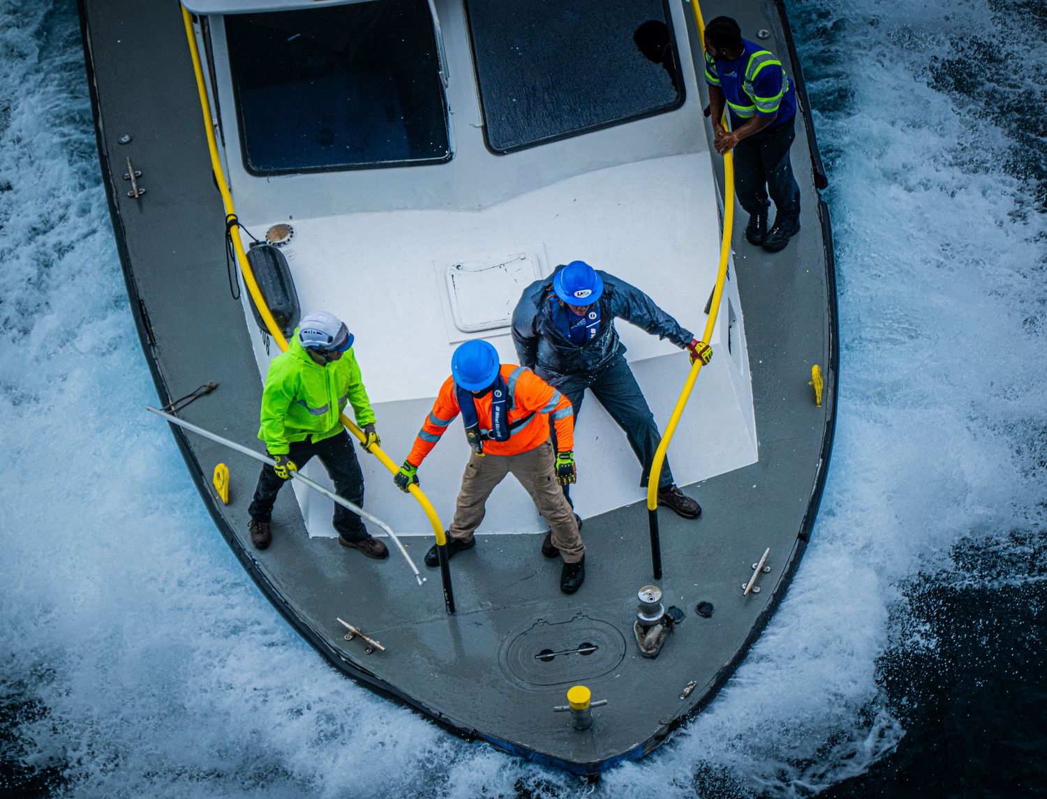 Aerial view of three workers in safety gear on a boat bow, navigating rough ocean waters.