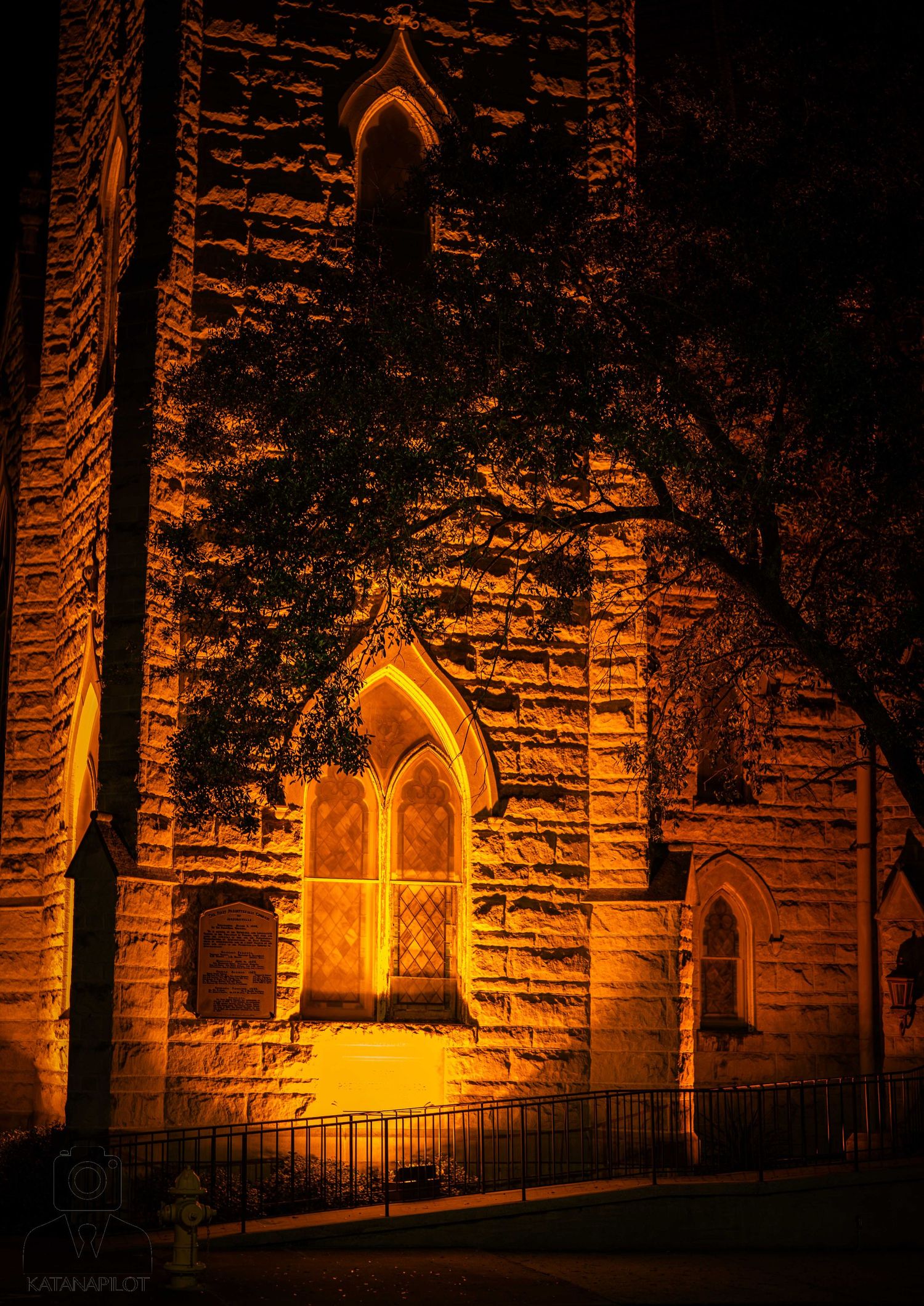 Gothic stone church tower illuminated by warm orange light at night, featuring arched windows and intricate stonework.