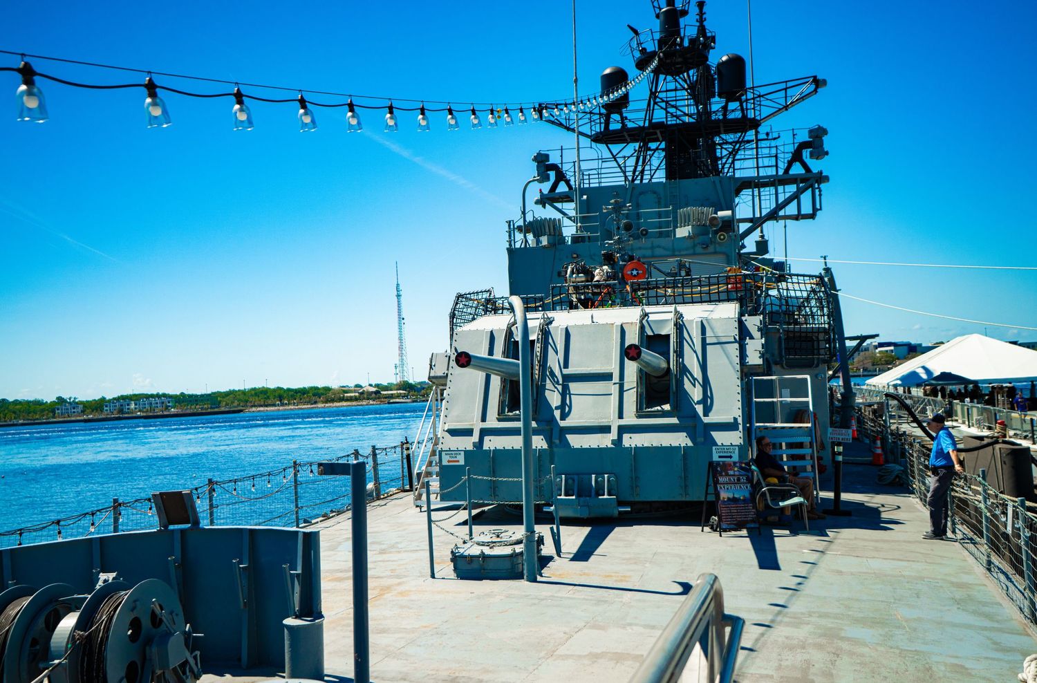 Navy warship deck with military superstructure, radar equipment, and blue sky over calm waterway on a sunny day.