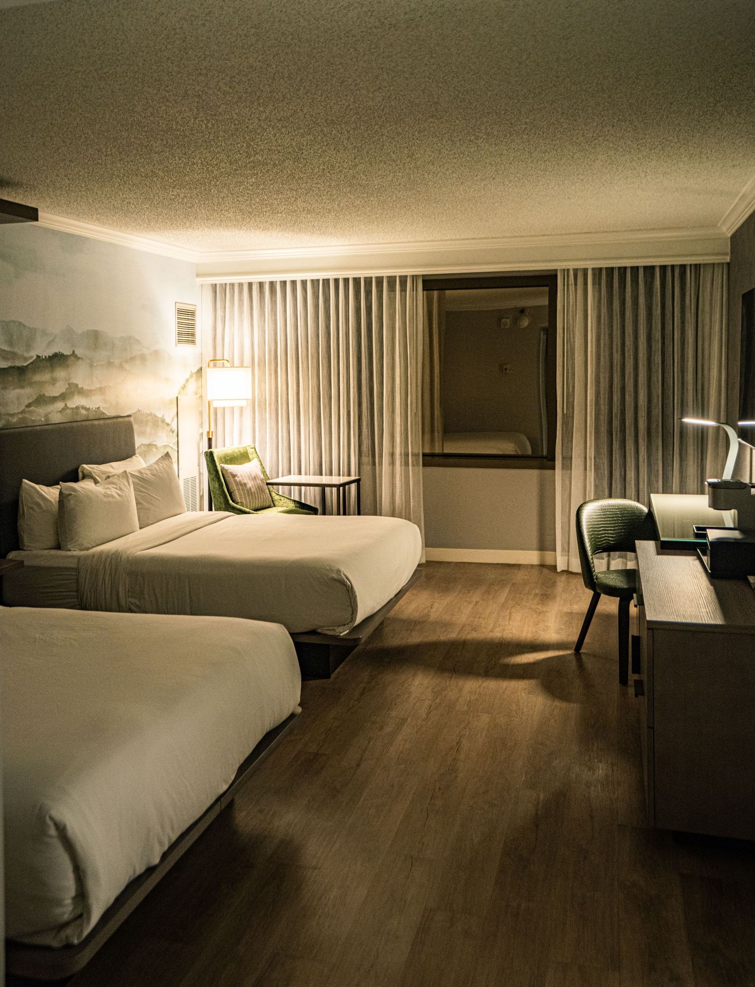 Modern hotel room with two queen beds, hardwood floors, neutral decor, desk, and elegant lighting.
