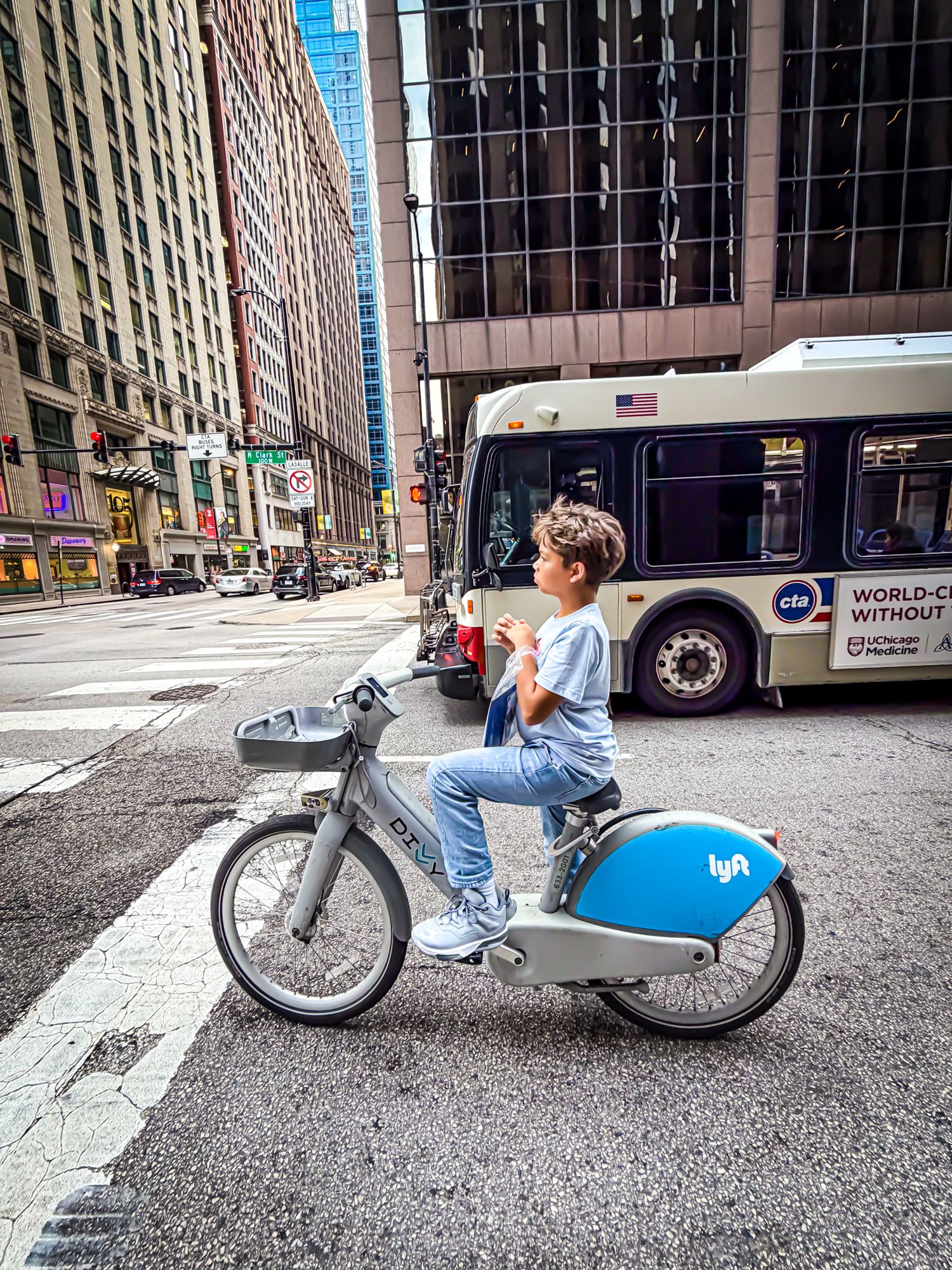 Child sitting on a Lyft bike-share scooter on a city street with a CTA bus and tall buildings in Chicago.