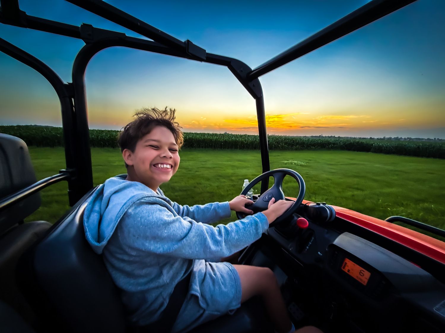 Smiling child driving a red off-road buggy at sunset with green fields and colorful sky in the background.
