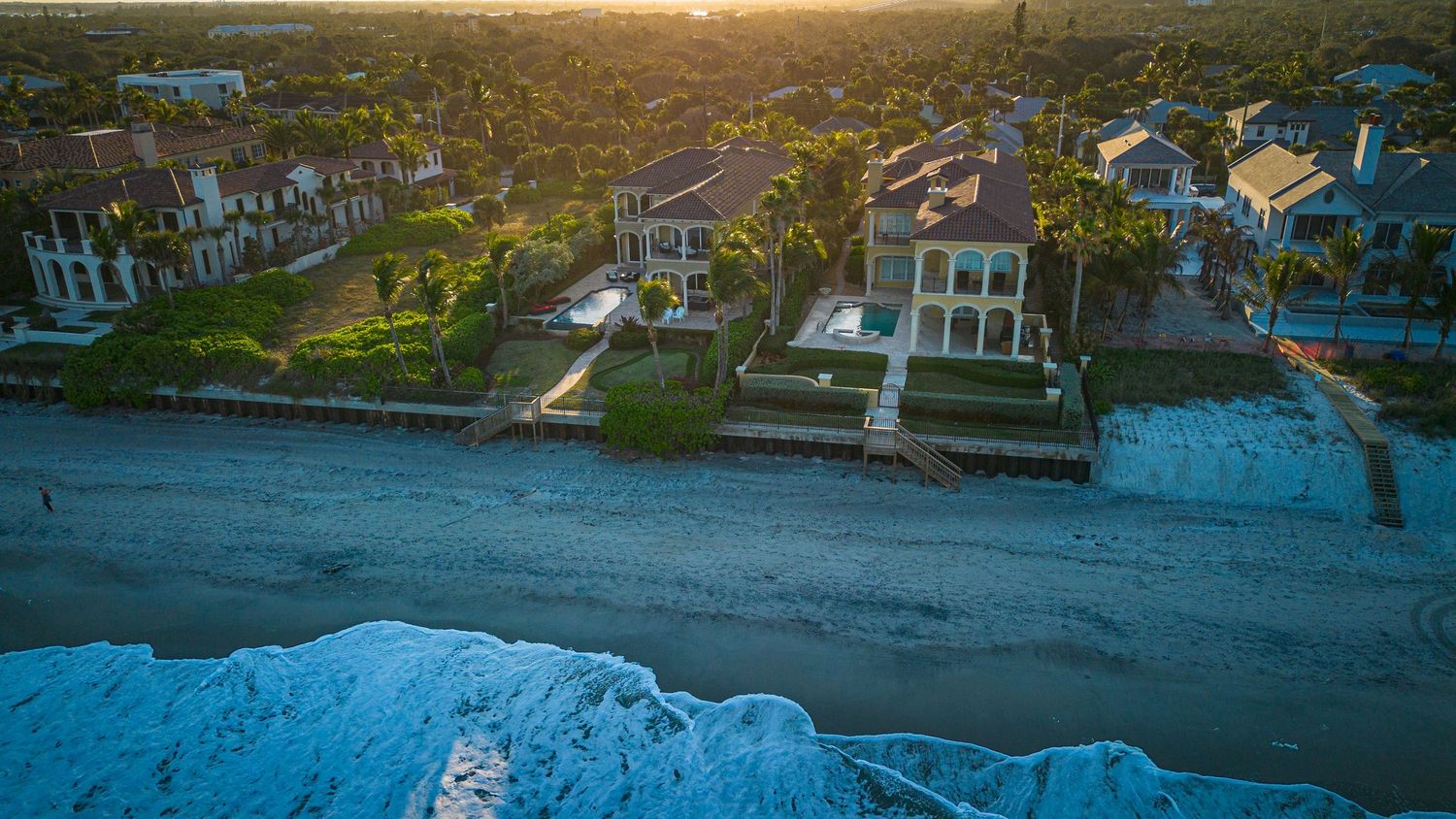 Sunset aerial view of waterfront homes along a dark sandy beach with rolling waves.