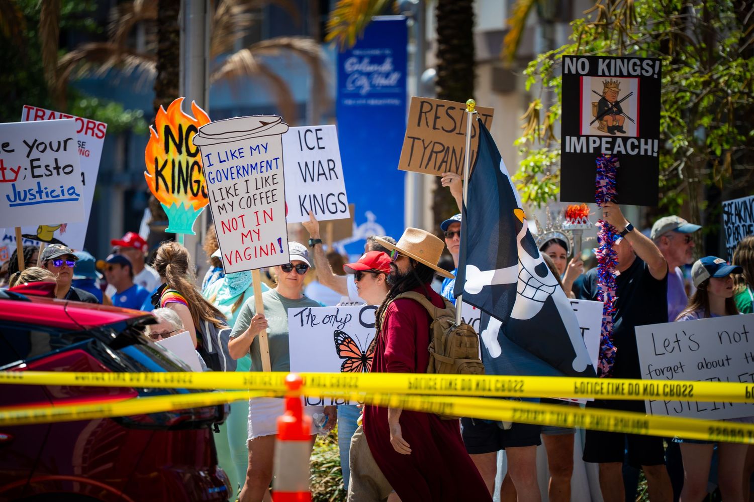 Protesters hold various signs at a political demonstration behind yellow caution tape in an outdoor setting.