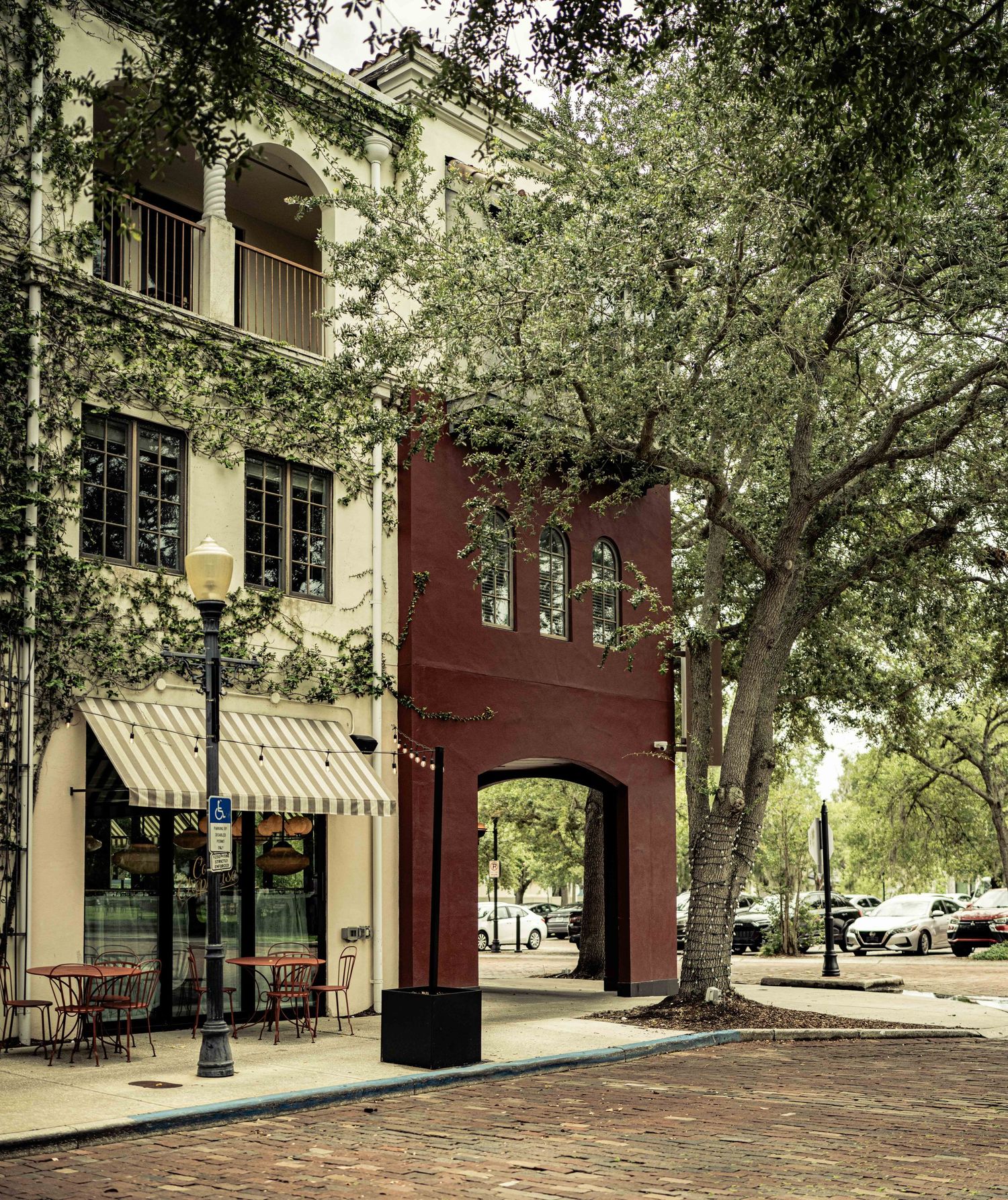 Historic brick building with arched gateway, tree-lined cobblestone street, and outdoor cafe seating in a charming Southern town square.