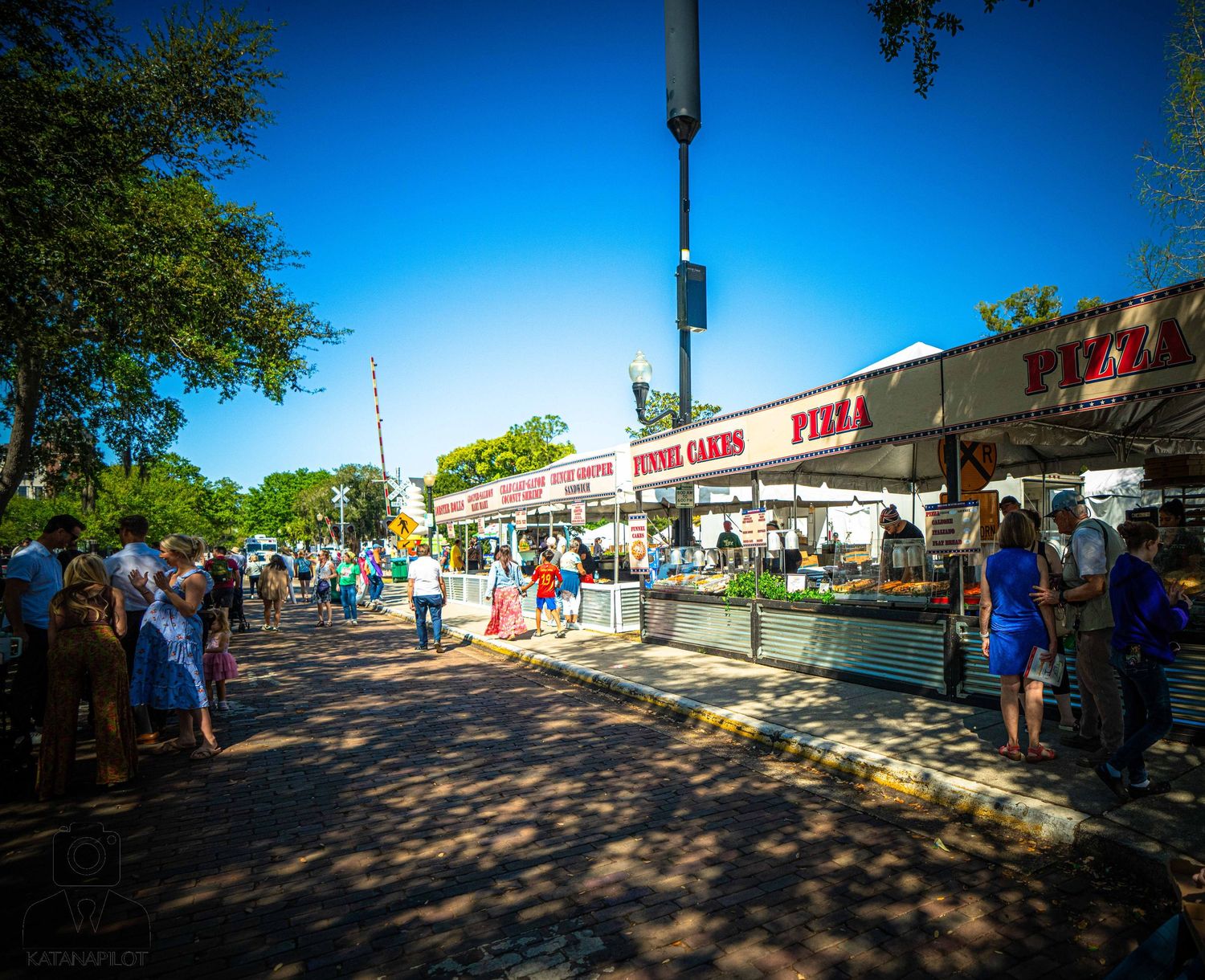 A row of white vendor tents lines a tree-shaded walkway at an outdoor market with visitors browsing.