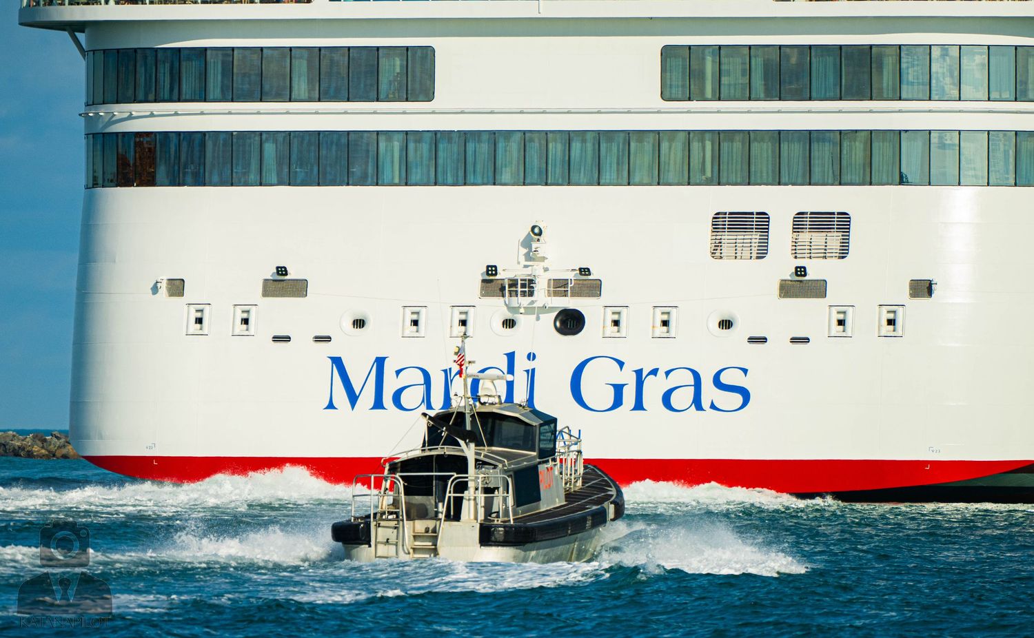 Tugboat sprays water while maneuvering alongside a large red and white vessel in calm waters.