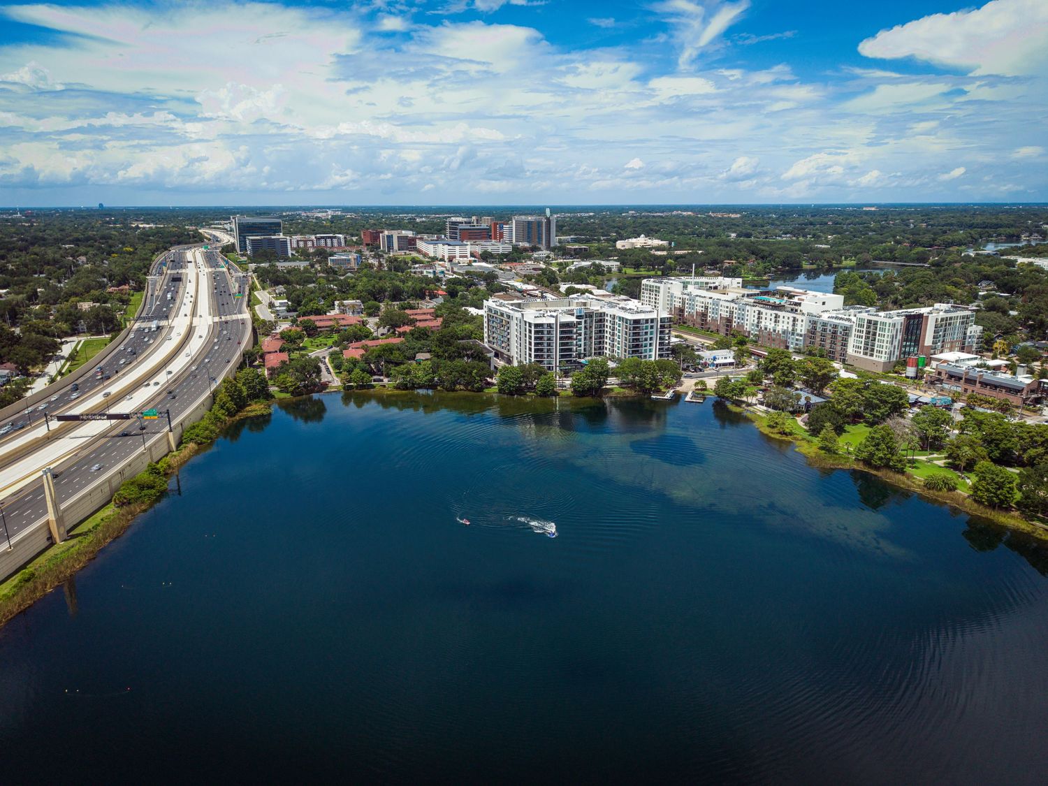 Aerial view of a Florida city skyline with a calm blue lake, a small boat, and a highway surrounded by lush green trees.