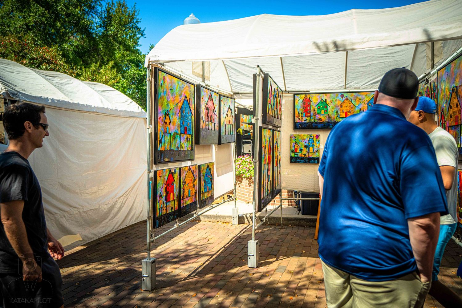 Row of white canopy tents line a sunny pathway at an outdoor art market with visitors browsing displays.