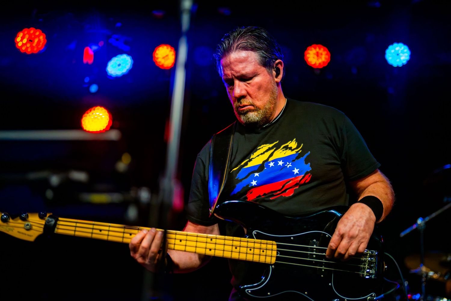 Musician playing bass guitar on stage under colorful lights, wearing a Venezuela flag t-shirt.