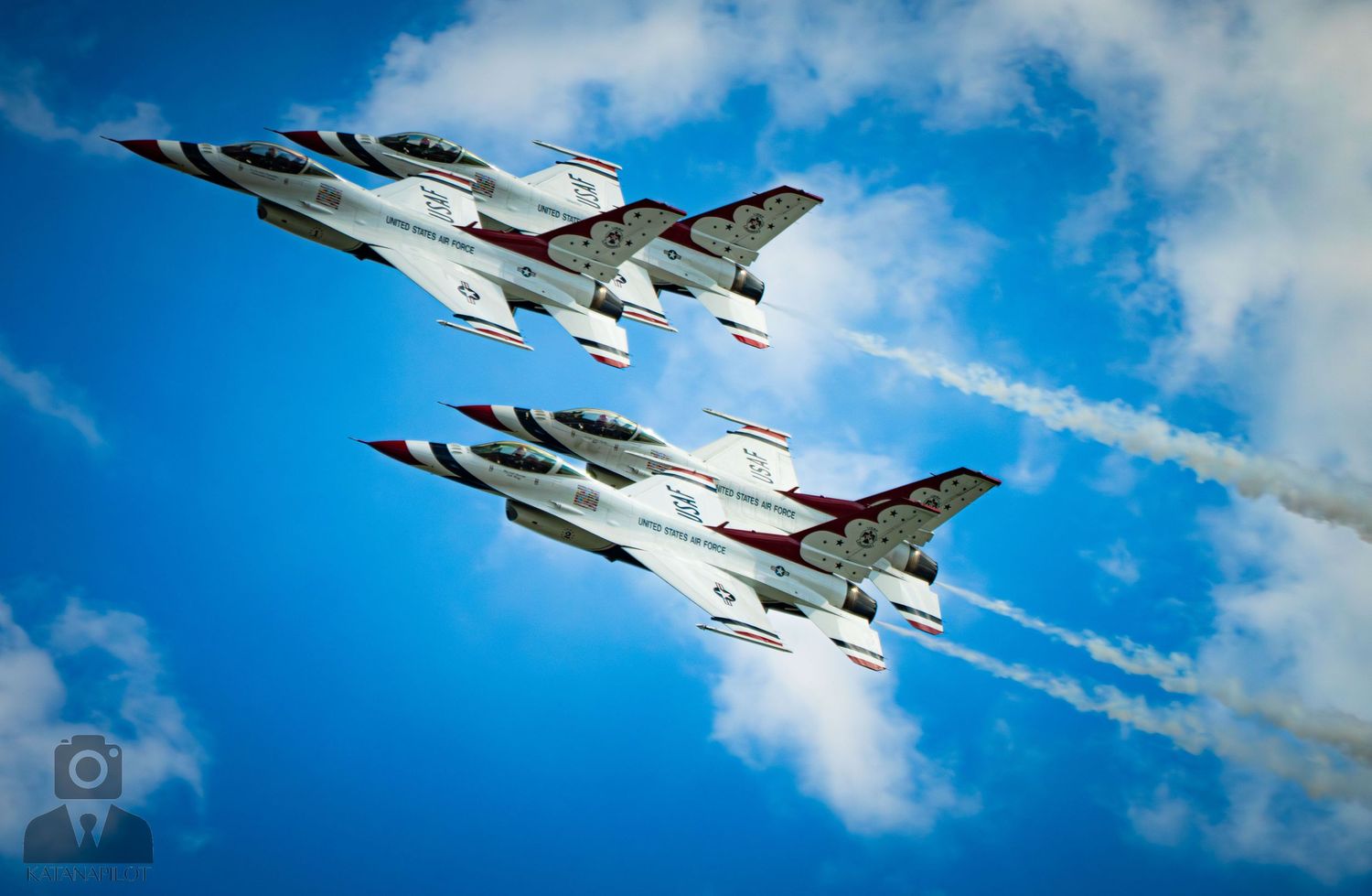 Two USAF Thunderbirds F-16 jets fly in close formation against a blue sky, leaving white smoke trails.