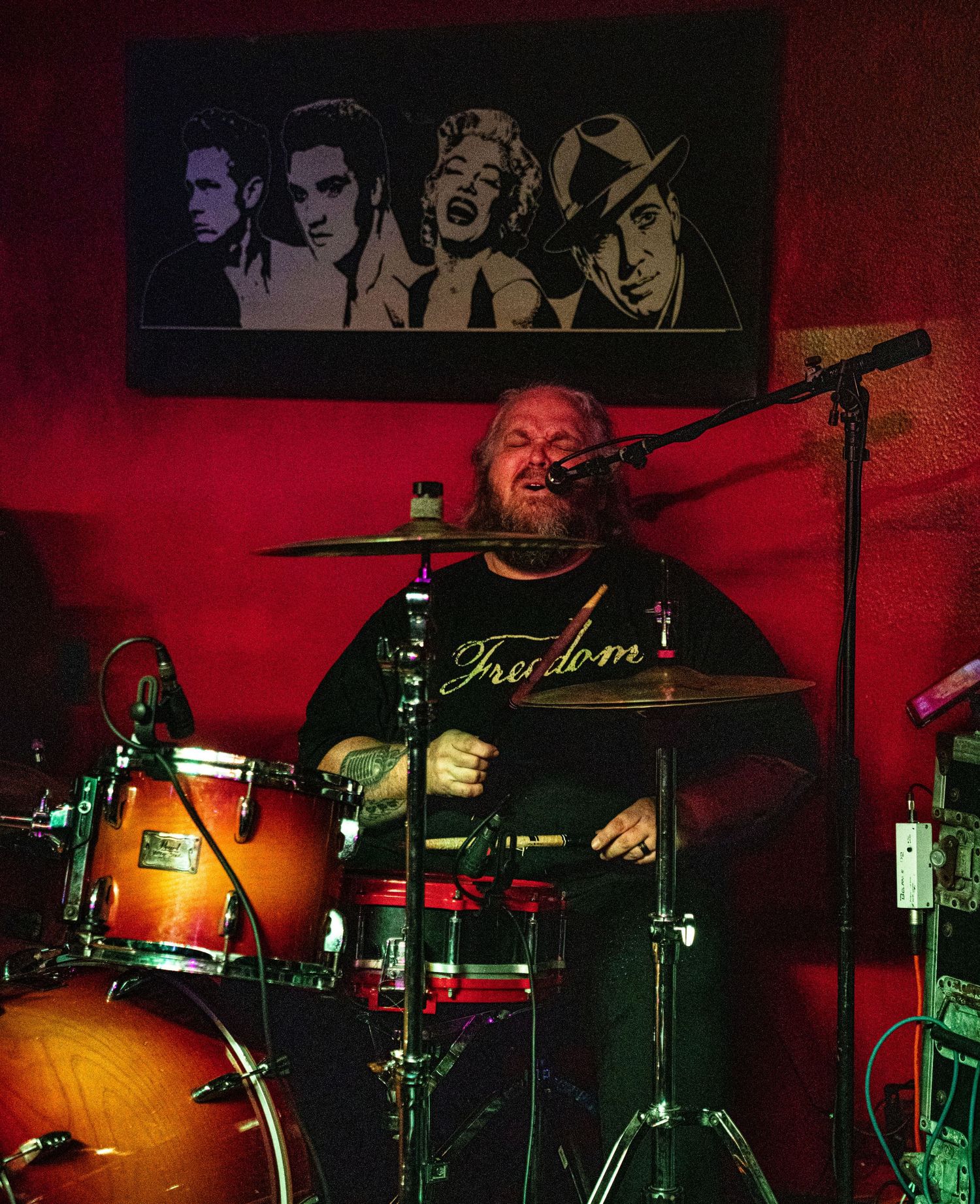 Drum kit and performer illuminated by red stage lighting with vintage portraits on the wall.