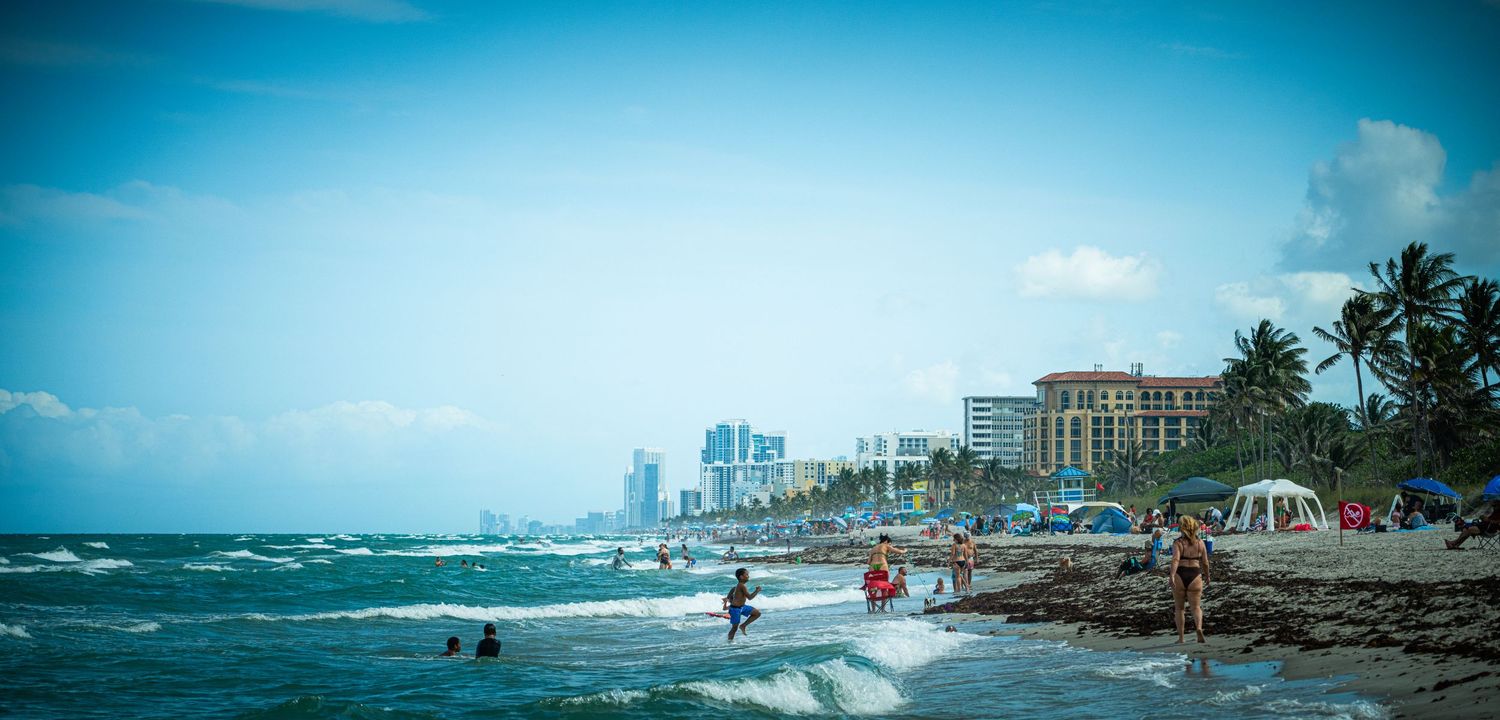 Coastal panorama showing beach, ocean waves, and high-rise buildings in the distance.