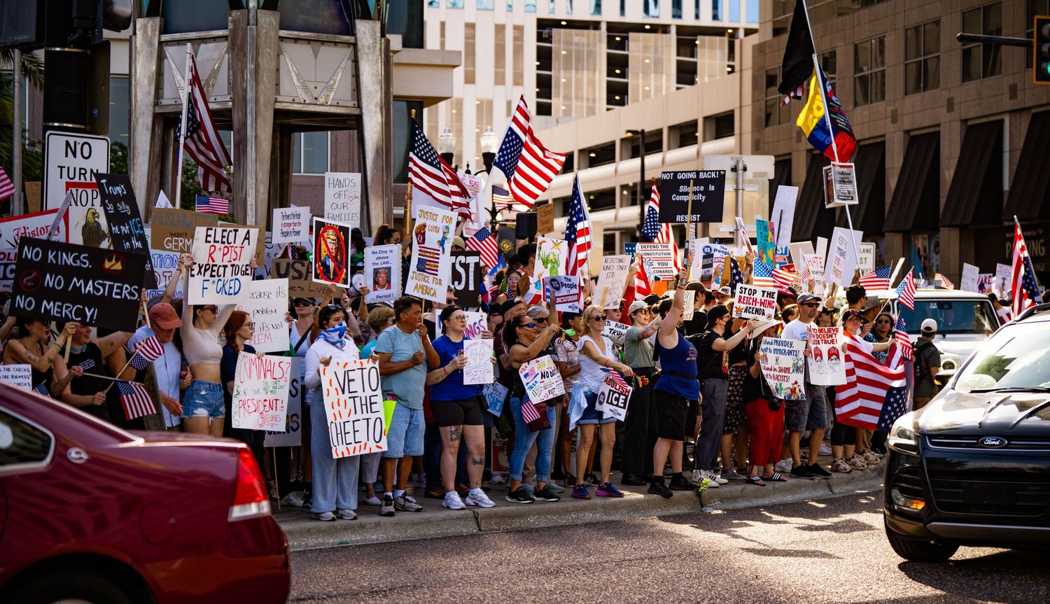 Crowd of demonstrators gathering on city street with protest signs and American flags.