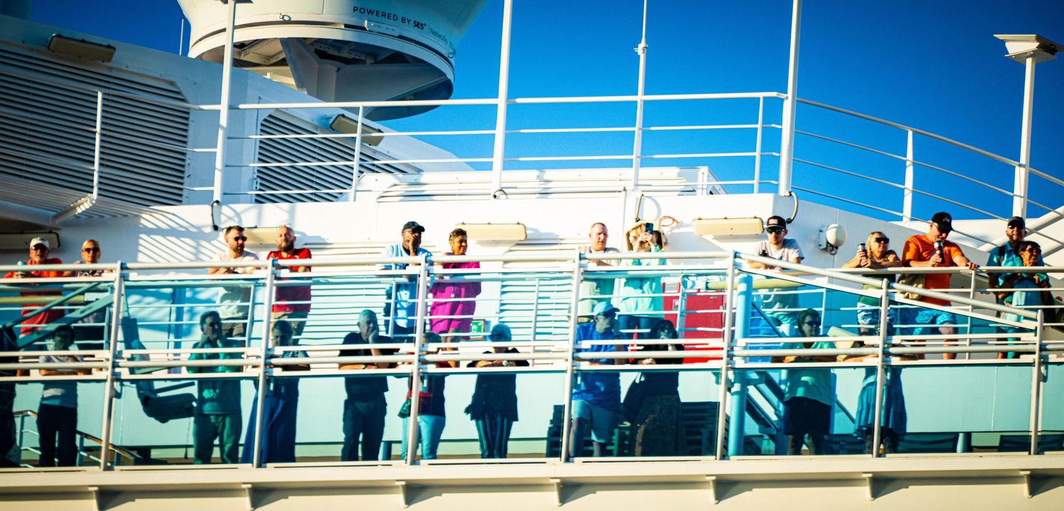 Cruise ship passengers enjoy views from multiple decks of a large ocean liner during a sunny day at sea.