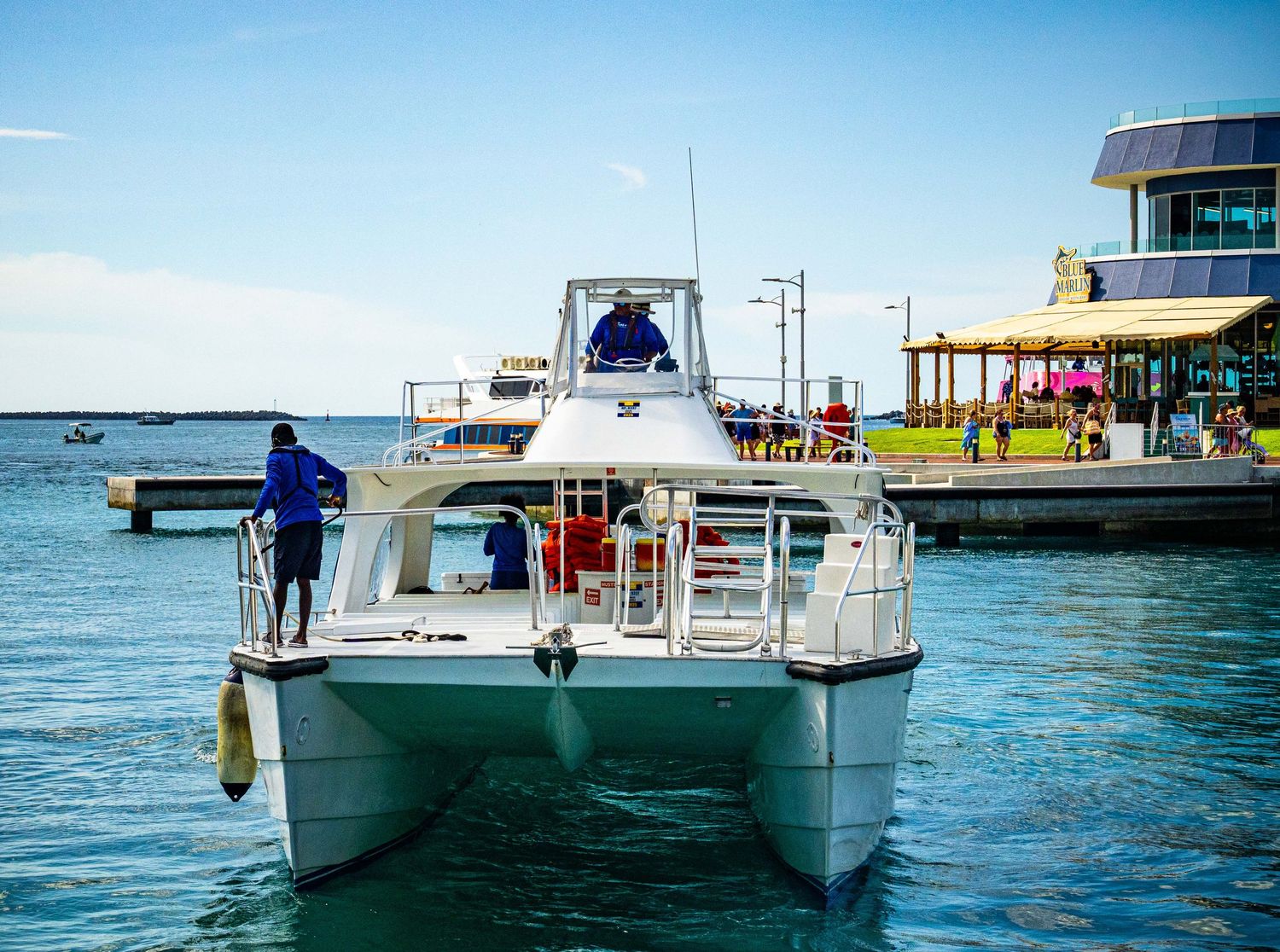 White catamaran boat docked at a marina pier on calm blue water with waterfront buildings in the background.