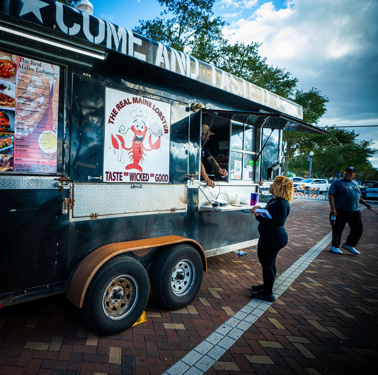 Black food truck parked on brick pavement with customers waiting outside under blue sky.