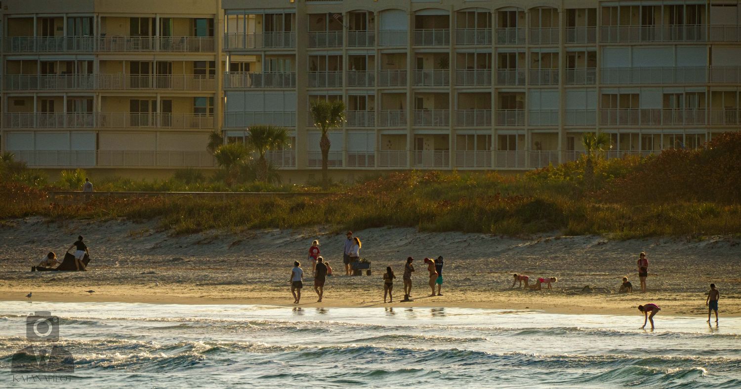 People enjoying a sunset beach walk along the shoreline with apartment buildings in the background during golden hour.