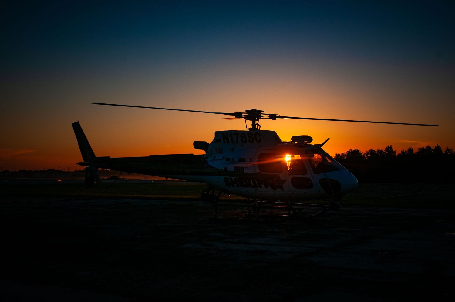 Helicopter silhouettes against dramatic orange sunset on airfield.
