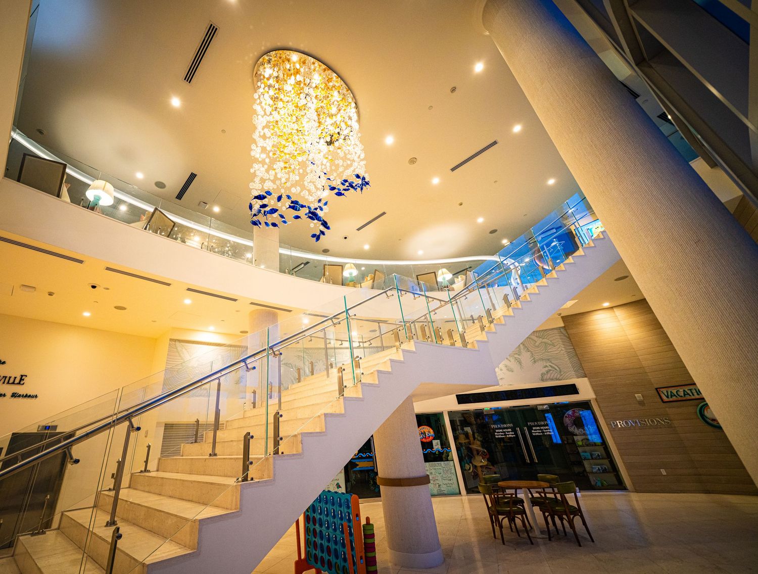 Elegant hotel lobby featuring a grand staircase with glass railings and a stunning blue chandelier in a bright atrium.