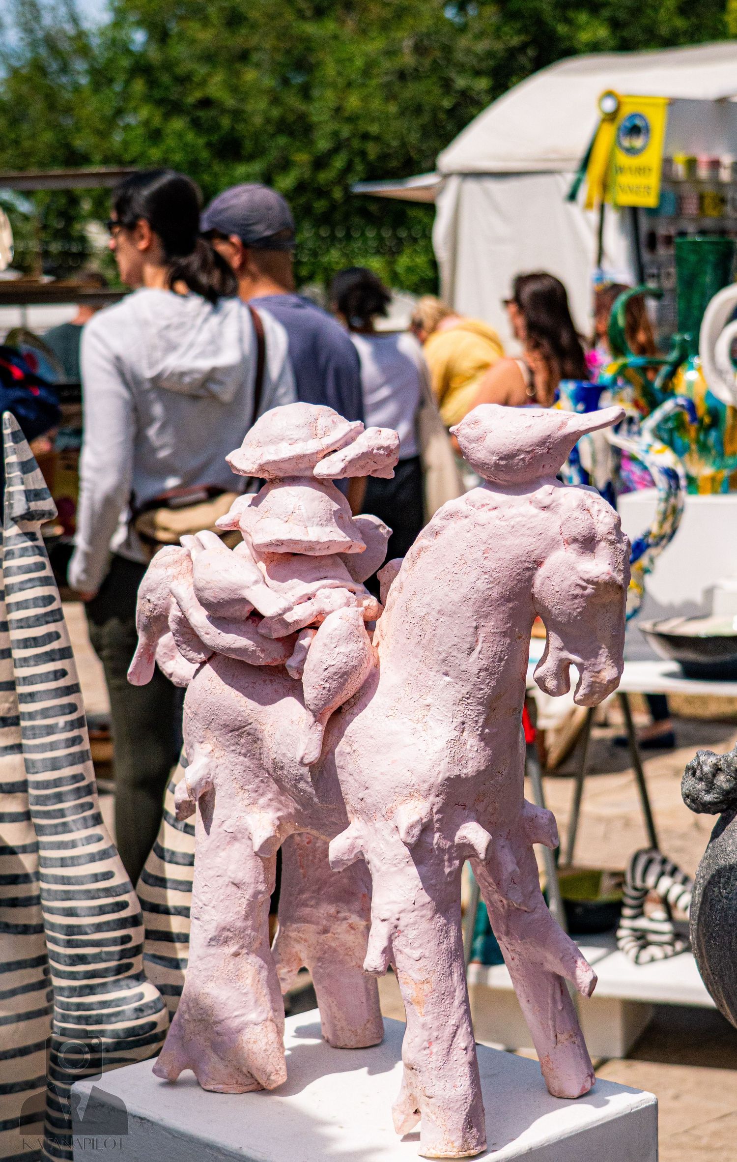 Pink sculptural figures displayed on pedestals at an outdoor art market.