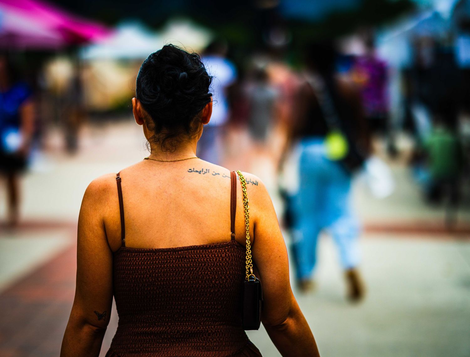 Woman with back tattoo and brown dress walks through a busy outdoor pedestrian area with blurred crowd.