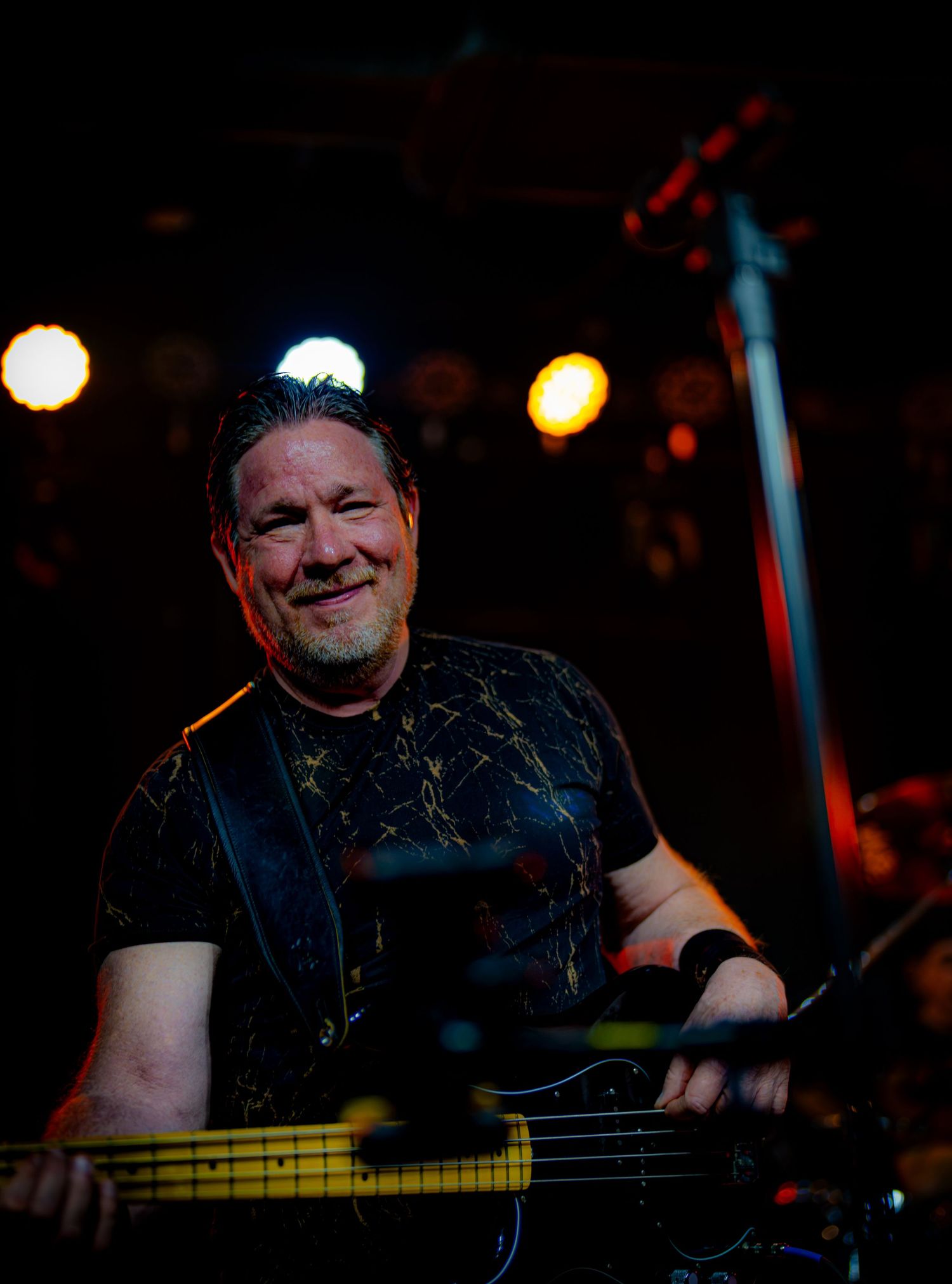 Smiling male bassist performing on stage under colorful concert lights in a dark venue.