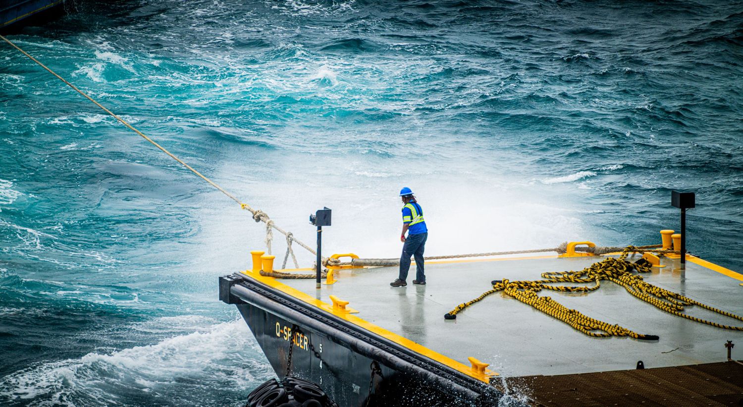 Sailor in blue hardhat managing ropes on ship deck amid rough turquoise ocean waves.