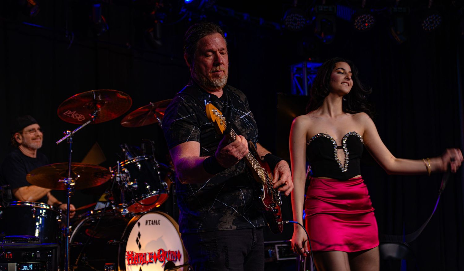 Band performing live on stage with guitarist and female singer in pink skirt under blue stage lights.