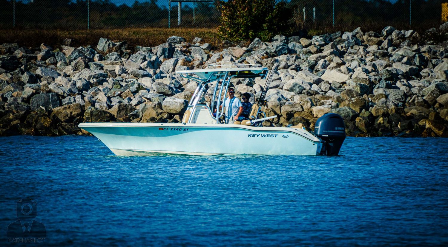 A scenic waterfront view showing boats and rocky breakwater along a calm blue shoreline.
