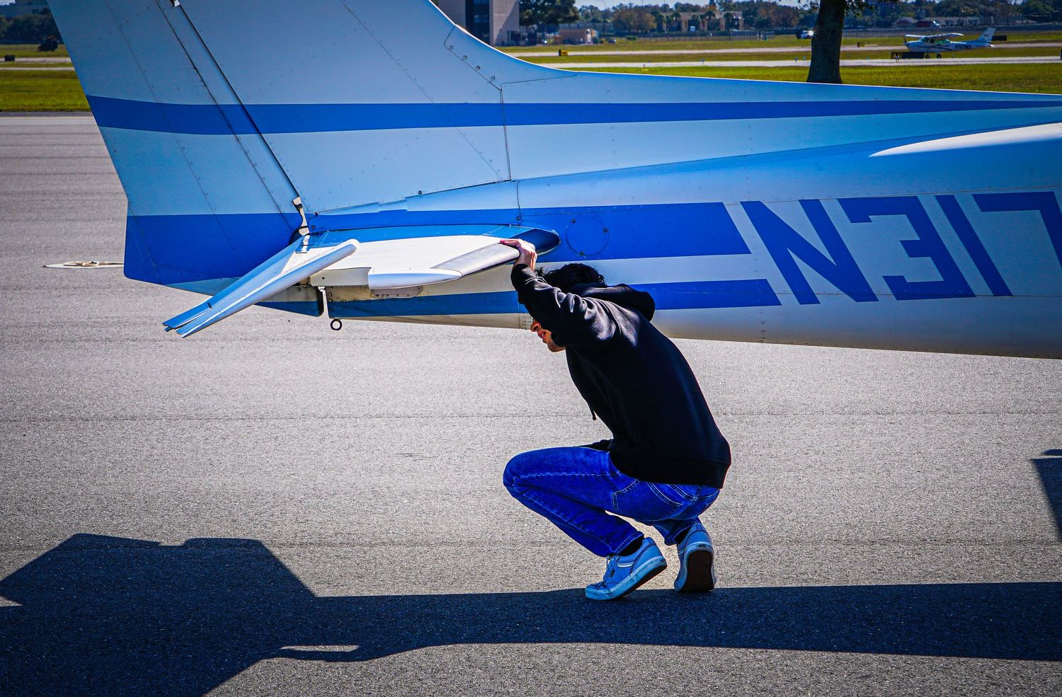 Person in blue crouching to inspect the wing of a small aircraft on an airport tarmac.