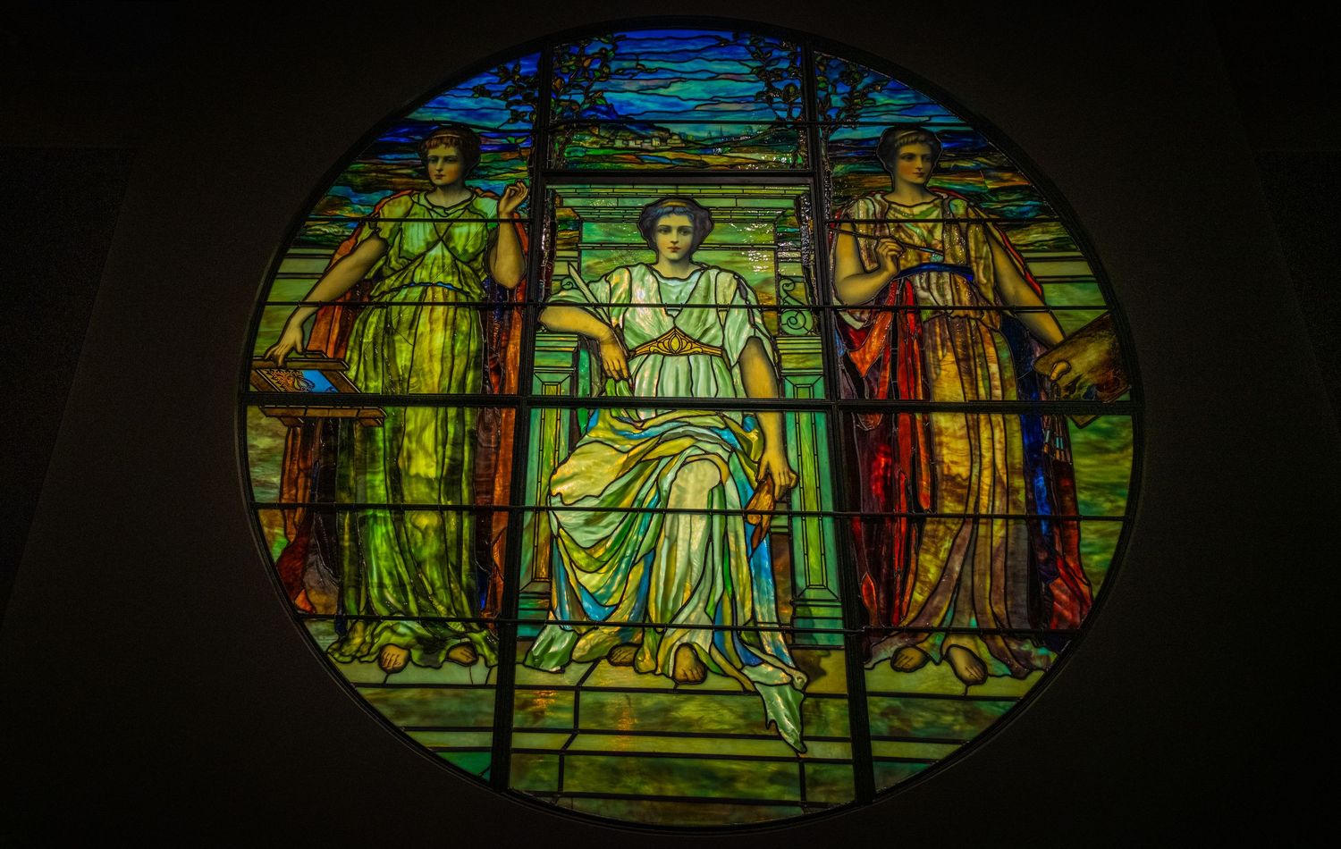 Circular stained glass window depicting religious figures in green, blue and red robes in a classical style.