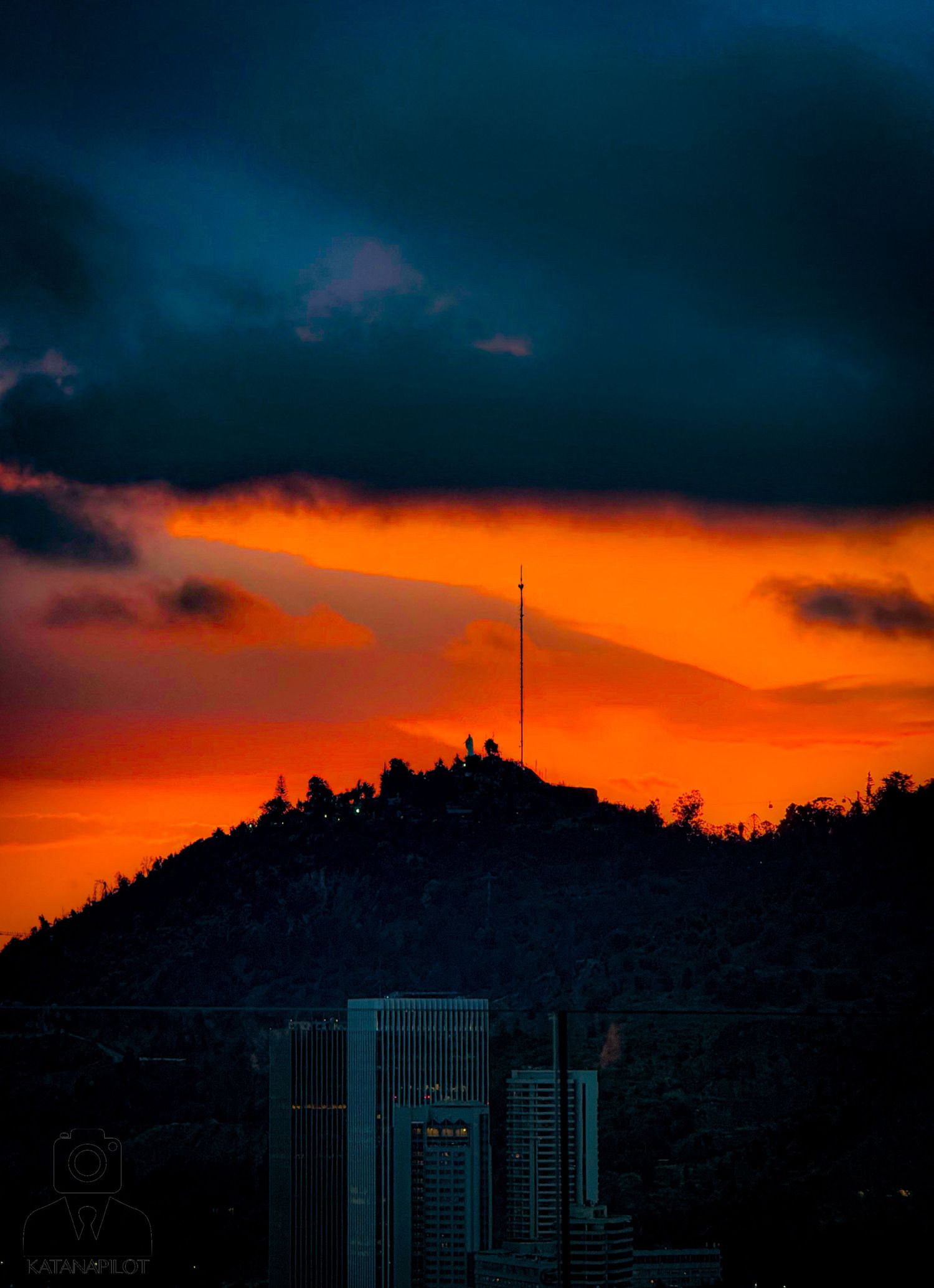 Dramatic orange sunset silhouettes city buildings against dark stormy clouds.