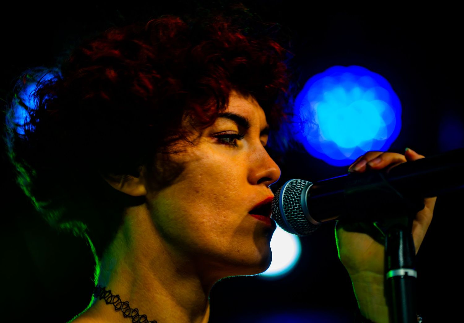 Close-up of female singer with curly red hair performing passionately into a microphone on stage.