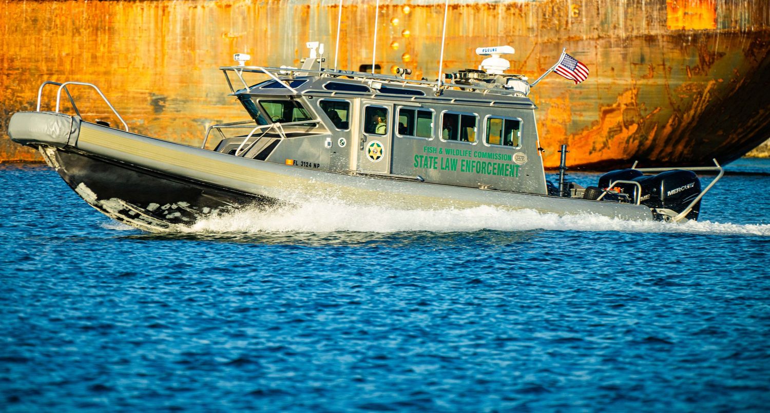 A military patrol boat speeds across blue waters against an orange industrial backdrop.