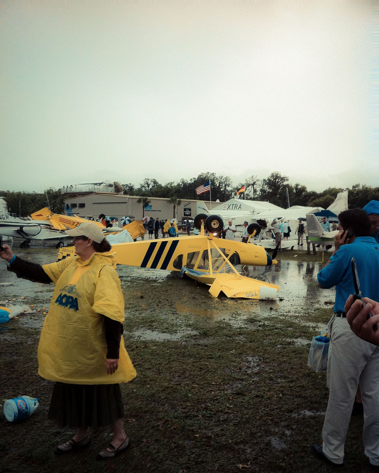Debris and damage from a tornado's aftermath with yellow emergency vehicles and response teams visible in the muddy field.