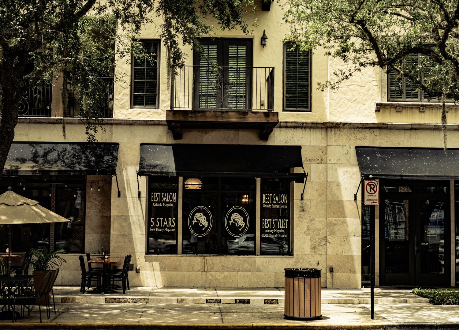 Quaint storefront with dark awnings, outdoor seating, and lush trees on a sunny urban street corner.