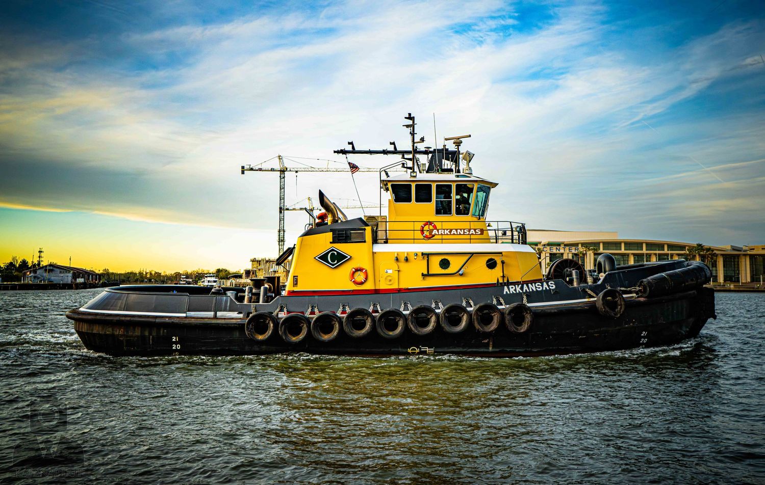 A large yellow industrial tugboat navigates across calm waters under a dramatic blue and cloudy sky.