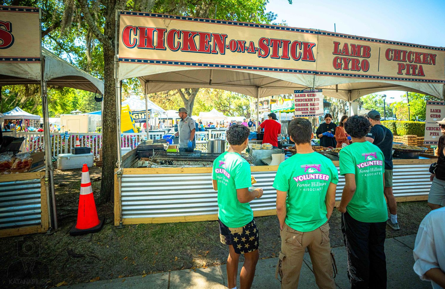 People in green shirts waiting in line at a chicken on a stick food vendor stand at an outdoor event.