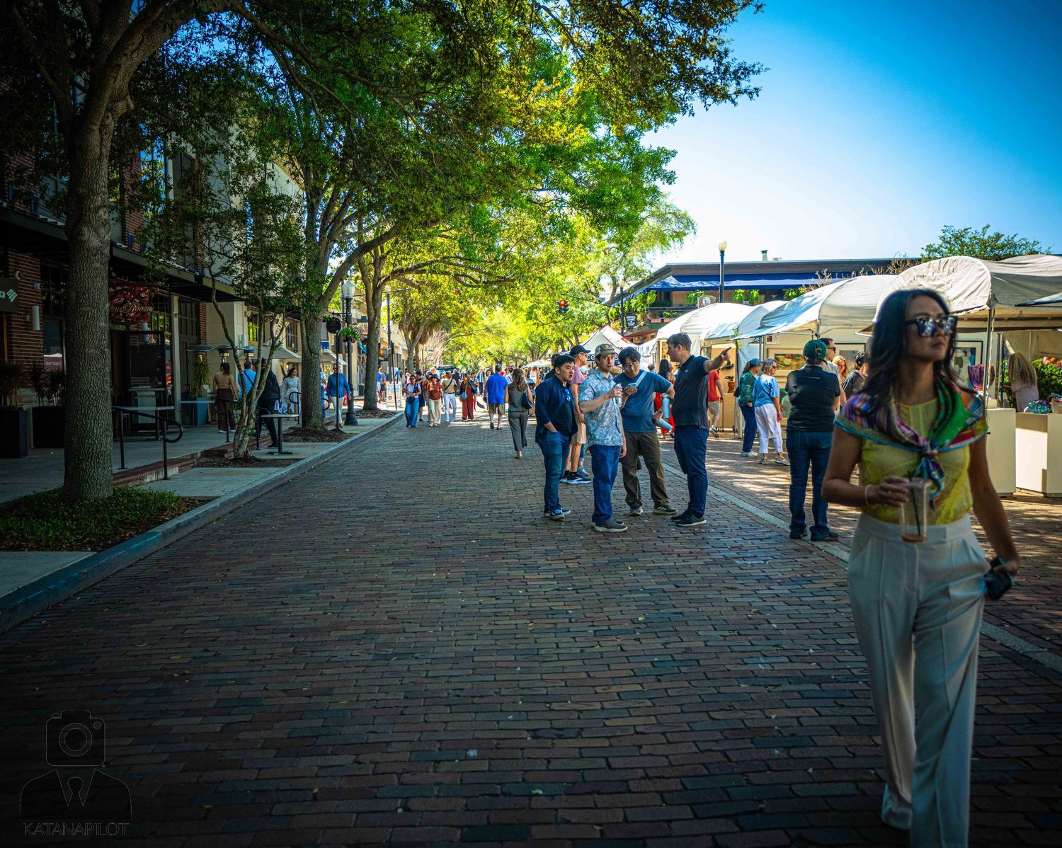 People browse outdoor market stalls along a brick-paved street lined with trees on a sunny day.