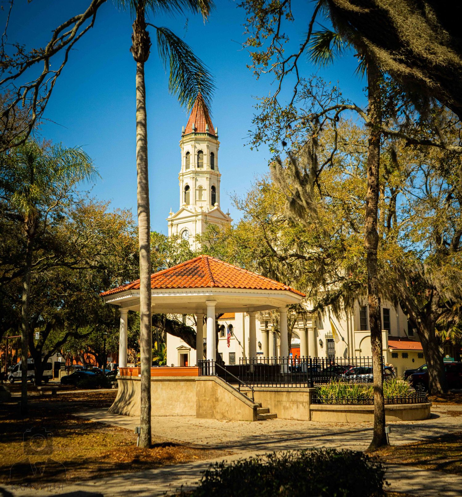 Historic Spanish-style church building with orange tile roof surrounded by moss-draped oak trees in a park setting.