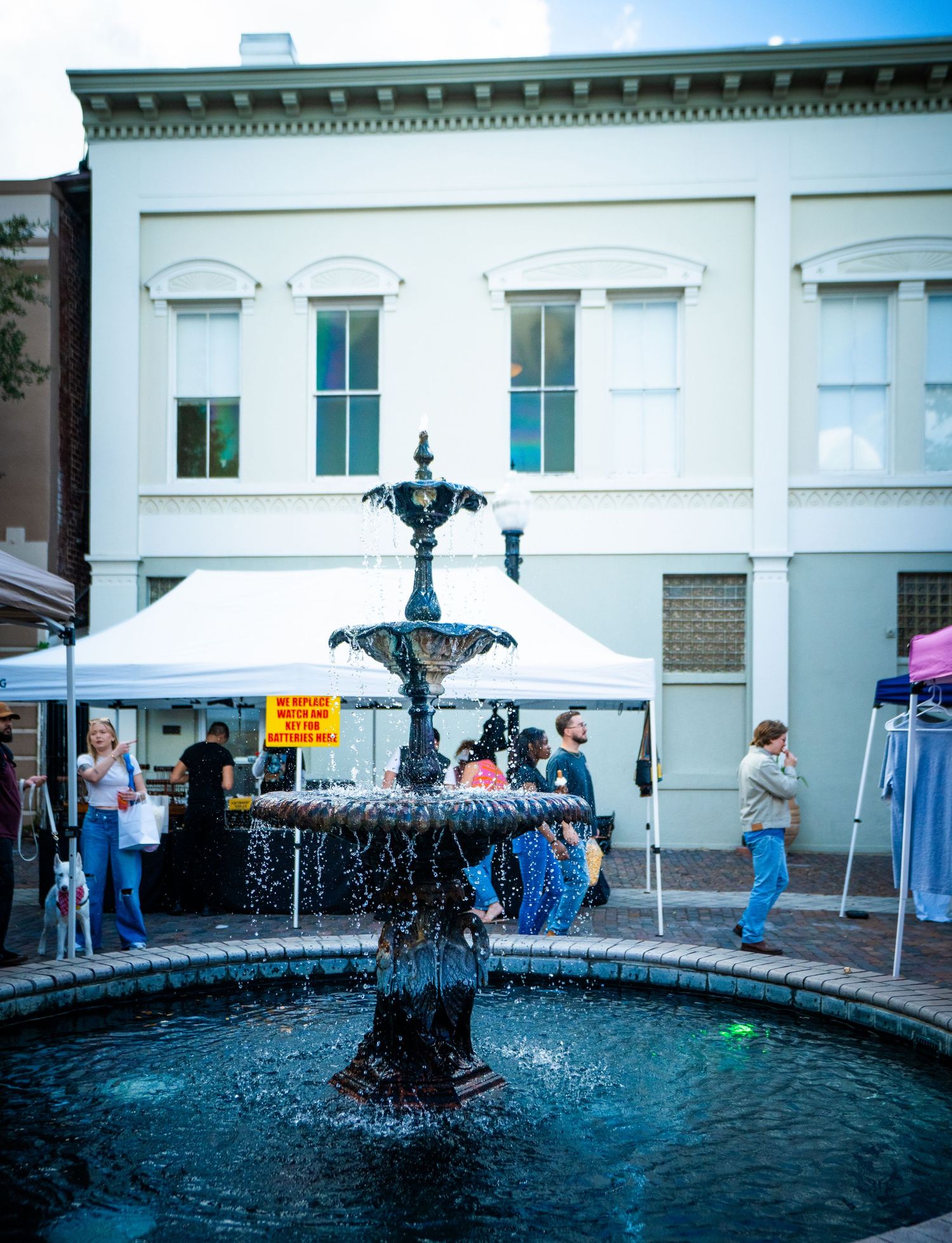 A decorative water fountain flows in front of white historic buildings with vendor tents and visitors at an outdoor market.
