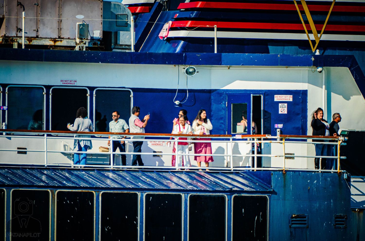Passengers gathered on the deck of a blue and white cruise ship during daytime sailing.