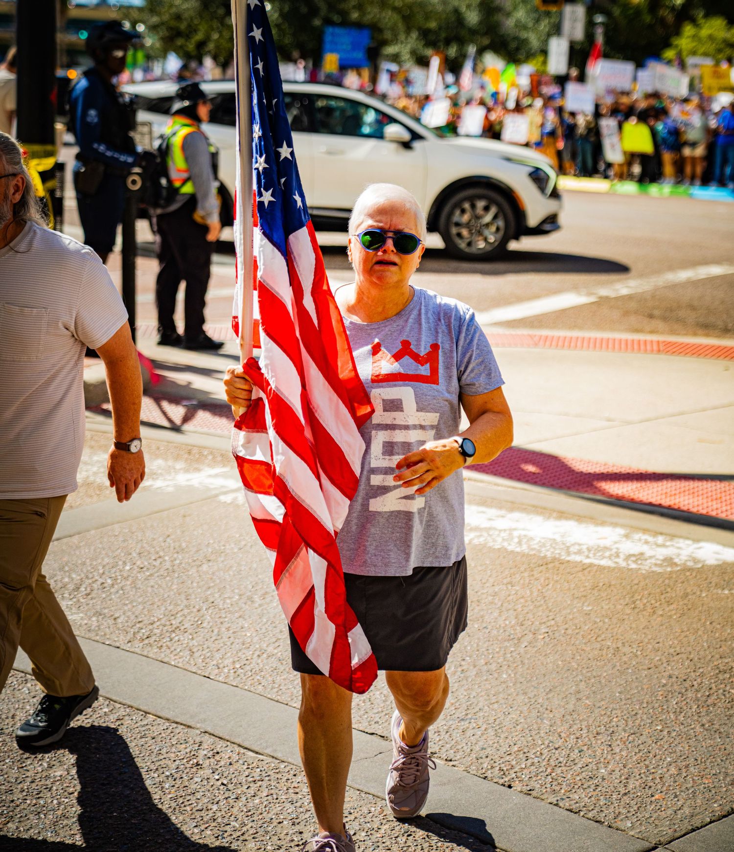 Demonstrators carrying American flags march along a sunny street during a protest.