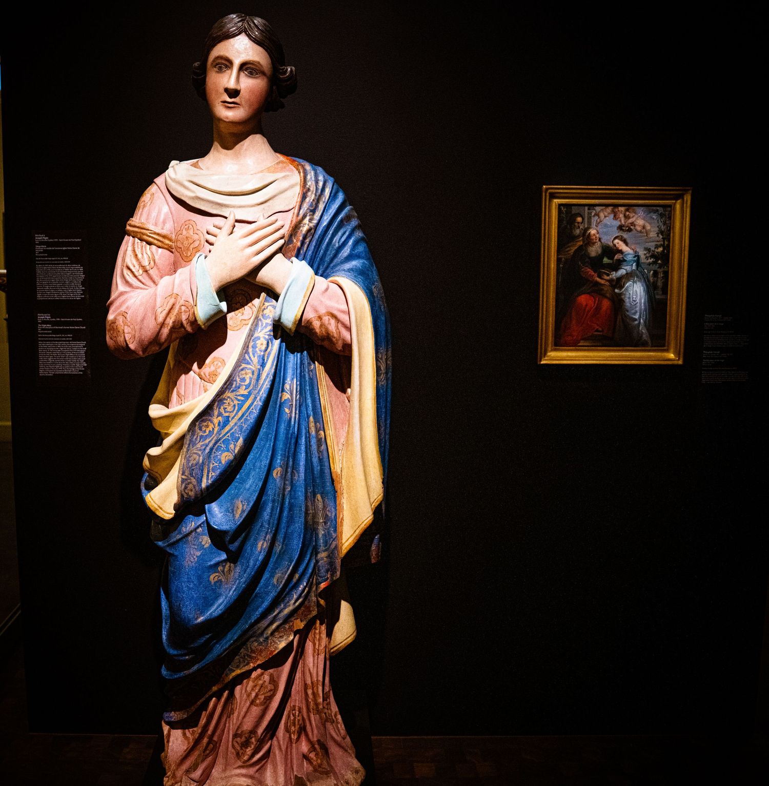 Classical statue in blue and pink robes displayed in dramatic lighting against dark background at museum.
