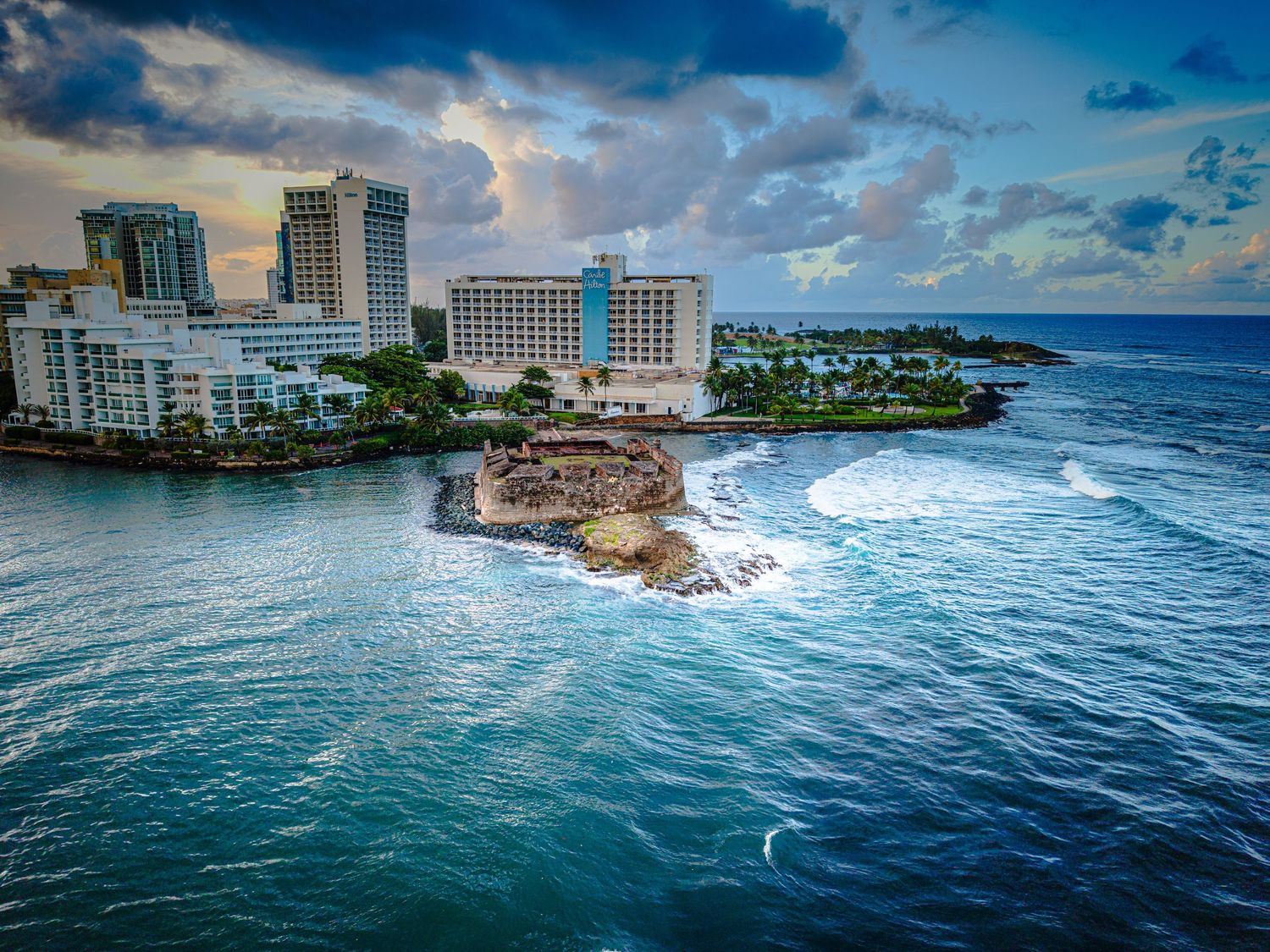 Dramatic oceanfront view of modern city skyline with turquoise waters and white-capped waves breaking on rocks.
