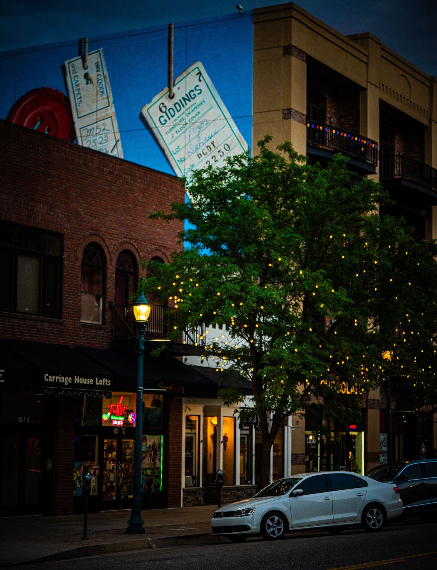 Charming downtown street at dusk with brick buildings, string lights, a glowing lamp post, and a white car parked along the sidewalk.
